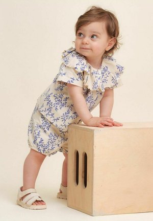 Blomstermønstret jumpsuit med flæse-besat halsudskæring, cremefarvet stof med blå blomster, parret med lysebrune sandaler, barn læner sig op ad en træboks.