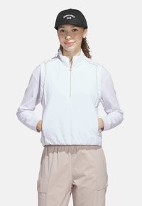 Hvid zip-up jakke med en struktureret krop, lange mesh ærmer og kontrasterende sømme. Båret med lyse beige bukser og en sort cap med hvid tekst.