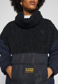 Svart fleece-pullover med hög krage, texturerad tyg och framficka. Har G-Star Originals-logotyp i gult på fickan.