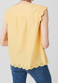 Geel mouwloos blouse met gegolfde randen en oogjesdetails. Lichte stof, achteraan een plooitje voor de pasvorm, gecombineerd met blauwe jeans.