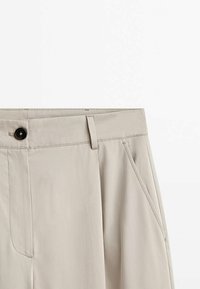 Beige broek met een vlakke voorkant, voorzien van een knoop- en ritssluiting, zijzakken en een zachte, stoffen textuur.