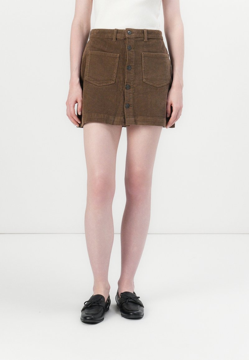 Femme portant une mini-jupe en velours côtelé marron avec des boutons sur le devant, de grandes poches avant, des chaussures noires sans lacets, et un haut blanc sans manches.