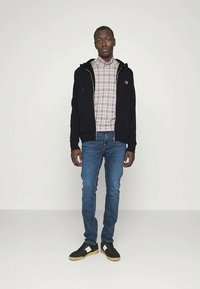 Čierna mikina na zips, károvaná košeľa, modré rifle a čierne tenisky s bielymi akcentmi, stojaci na jednofarebnom pozadí.