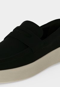 Μαύρο loafer slip-on με παχιά ανοιχτό μπεζ σόλα και φαρδιά λωρίδα στο μπροστινό πάνω μέρος σε απλό ανοιχτό φόντο.