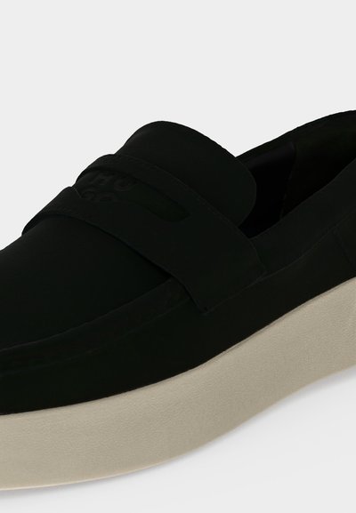Mocassin noir sans lacets avec semelle épaisse beige clair et large bride sur l’avant supérieur sur un fond clair uni.