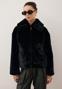 Kobieta w czarnej sztucznej futrze, zapinanej na zamek, z dużym kołnierzem, ciemnych okularach przeciwsłonecznych, złotych kolczykach, pierścionkach i ciemnoszarych spódnicach plisowanych, stojąca przy białej ścianie.