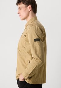 Veste kaki clair en tissu texturé, dotée d'une poche poitrine boutonnée, de poches latérales et d'un insigne sur la manche gauche.