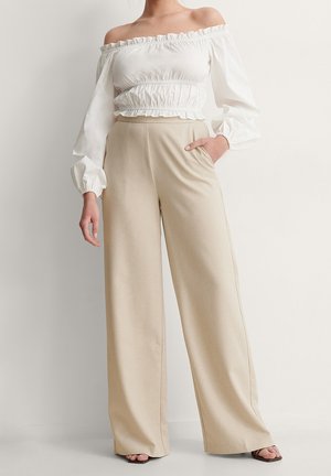 Vrouw draagt beige broek met wijde pijpen en een witte blouse met lange mouwen en blote schouders, staande met één hand in de zak.