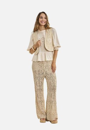 Mujer con chaleco beige bordado, blusa crema con mangas abullonadas y pantalones anchos de crochet, sonriendo y posando contra un fondo liso.