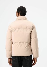 Beige donzen jas met hoge kraag, doorgestikt patroon en elastische manchetten; ontworpen voor warmte en comfort, geschikt voor casual gebruik.