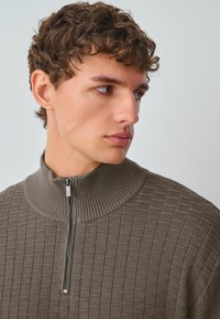 Brązowy sweter zapinany na zamek z teksturowanym wzorem w kratkę i ściągaczem przy kołnierzyku. Posiada krótki zamek przy szyi, wykonany z miękkiego materiału.
