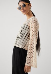 Pull en dentelle de couleur pêche clair et noir, avec des manches longues et un design cropped, associé à un pantalon noir et des lunettes de soleil.