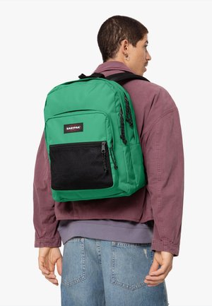 Mochila verde con bolsillo de malla negra, compartimentos con cremallera y correas ajustables, llevada sobre una camiseta gris y una chaqueta púrpura.