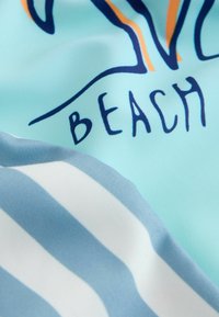 Tessuto azzurro chiaro con testo navy "BEACH" e parte di un disegno arancione e blu scuro sopra, accanto a un tessuto a righe bianche e blu.