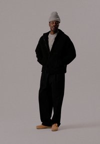 Black zip-up jacket, gray t-shirt, loose black trousers, and beige slip-on shoes. Gray knit beanie. Model stands against a neutral background.