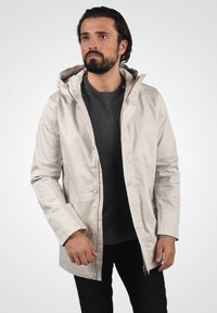 Beige vanntett jakke med glidelås foran og trykknapper, justerbar hette og lange ermer, iført over en grå genser.