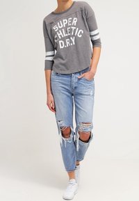 Grå kortärmad sweatshirt med vit text och ränder, i kombination med slitna ljusblå jeans. Modellen bär vita sneakers.