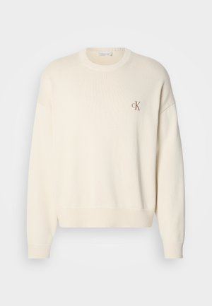 Suéter de punto color crema con textura acanalada, escote redondo y hombros caídos. Presenta un pequeño logotipo bordado en marrón en el pecho.