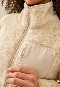 Beige Fleecejacke mit hohem Kragen, strukturiertem Stoff und einer kontrastierenden, glatten Tasche mit Reißverschluss an der Seite.