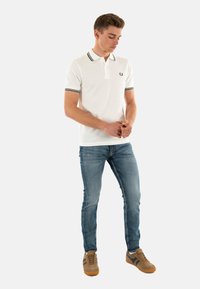 Polo shirt bianco con colletto a righe nere e verdi, abbinato a jeans azzurri e scarpe da ginnastica marroni con accenti blu scuro.