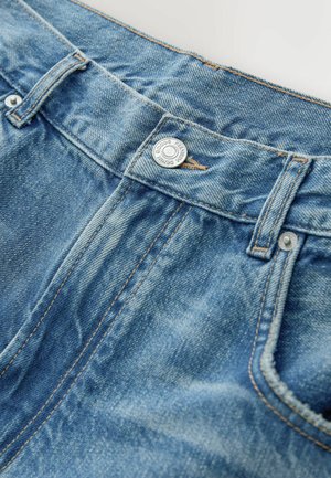 Vaaleansiniset farkut, joissa on klassinen viiden taskun malli. Niissä on hopeanvärinen napin suljin ja kontrastivärinen ruskea ommel.