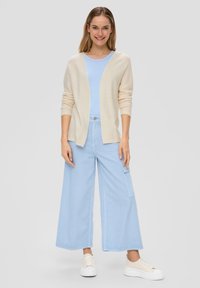 Licht beige cardigan over een lichtblauw T-shirt, gecombineerd met luchtige lichtblauwe broek met brede pijpen en witte veterschoenen. Casual, relaxed fit.