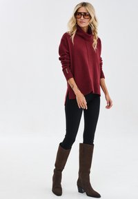 Bordowy sweter z golfem, ozdobiony prążkowanymi akcentami, w połączeniu z czarnymi legginsami i brązowymi kozakami do kolan. Gładka struktura, luźny fason.