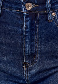 Jeans de mezclilla azul oscuro con una textura suave, que cuentan con un cierre de botón frontal, costuras doradas y un diseño estándar de cinco bolsillos.