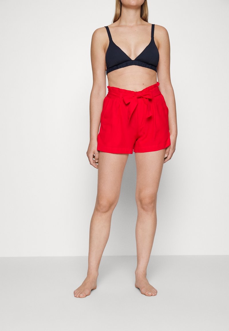 Femme portant un bralette noir et un short rouge taille haute avec une ceinture nouée, se tenant pieds nus devant un fond blanc uni.