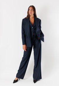 Completo blu con righe sottili composto da un blazer su misura, un gilet aderente e pantaloni a gamba larga. Tessuto liscio con accenti sottili. Tacchi alti neri a punta.