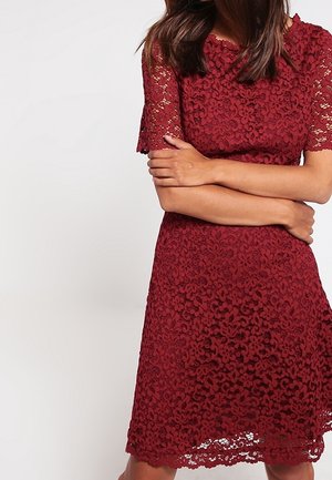 Kvinde iført knælange, kortærmede bordeauxfarvede blonderkjole, armene krydsede, med løst hår, på ensartet lysegrå baggrund.