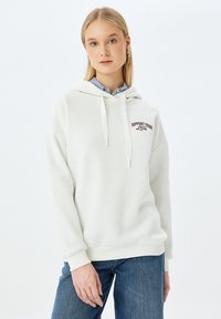 Sudadera blanca con bolsillo canguro y cordón. Presenta el texto "APOYA A TU LOCAL" en marrón oscuro. Combinada con una camisa a rayas azul y jeans azules.