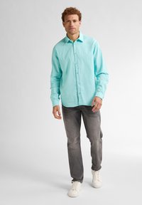 Camicia azzurra con bottoni, maniche lunghe, tessuto leggero. Indossata con jeans grigi e sneakers bianche. Il modello è in posizione eretta su uno sfondo chiaro.