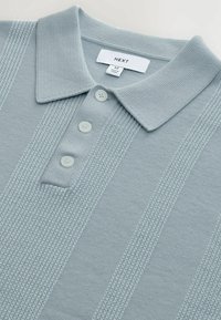 Lichtblauwe poloshirt van gebreide stof, met een gestructureerd streepjespatroon, een kraag en drie witte knopen. Etiket zichtbaar aan de binnenkant.