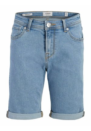 Garçon debout, une main dans la poche, portant un t-shirt blanc avec un motif bleu, un short en jean roulé, des chaussettes blanches rayées et des baskets noires.