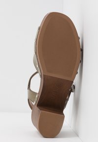 Sandale avec une semelle en caoutchouc texturée marron et un petit talon, avec des clous métalliques le long des côtés et une bride réglable.