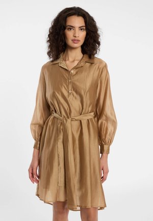 Femme aux cheveux bouclés et foncés portant une robe-chemise ceinturée marron clair brillante à manches longues et col, debout devant un fond blanc.