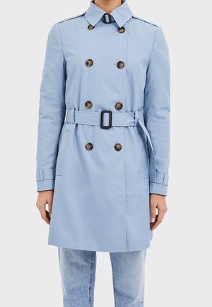 Vrouw draagt een lichtblauwe dubbelgeknoopte trenchcoat met riem, gecombineerd met lichtblauwe jeans, staand tegen een effen achtergrond.