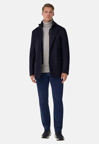 Granatowa marynarka z dwoma przednimi kieszeniami, nałożona na jasnoszary sweter z golfem. Do tego ciemnoniebieskie spodnie i czarne trampki.