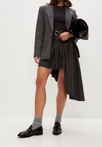 Grau maßgeschneiderte Blazer über einem schwarzen Oberteil, kombiniert mit einem hochsitztenden Rock mit asymmetrischem Saum. Abgerundet mit schwarzen Loafers und grauen Socken.