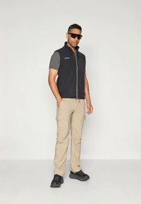 Melns bezpiedurkņu žakets virs pelēka krekla, apvienots ar bēšiem cargo biksēm un melnām kurpēm. Ievērības cienīgas detaļas ietver saulesbrilles un vairākas kabatas.