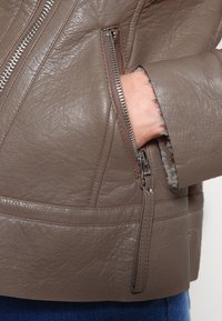 Veste en cuir marron présentant une finition texturée, des zips marquants et un détail de poche près du poignet avec une doublure douce.