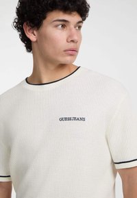 Baltos tekstūruotas trumpomis rankovėmis marškinėliai su juodu apvadu ant apykaklės ir rankovių, su mėlynu siuvinėtų "GUESS JEANS" logotipu ant krūtinės.