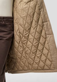 Μπεζ επενδεδυμένο μπουφάν με διαμαντένιο σχέδιο, με λεία υφή και στρογγυλή ένωση. Συνδυασμένο με σκούρα καφέ παντελόνια.