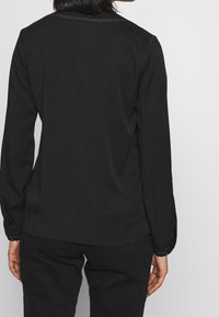 Maglietta nera a maniche lunghe con design semplice, morbido al tatto e dettagli di cucitura sottili al colletto, indossata con pantaloni neri.