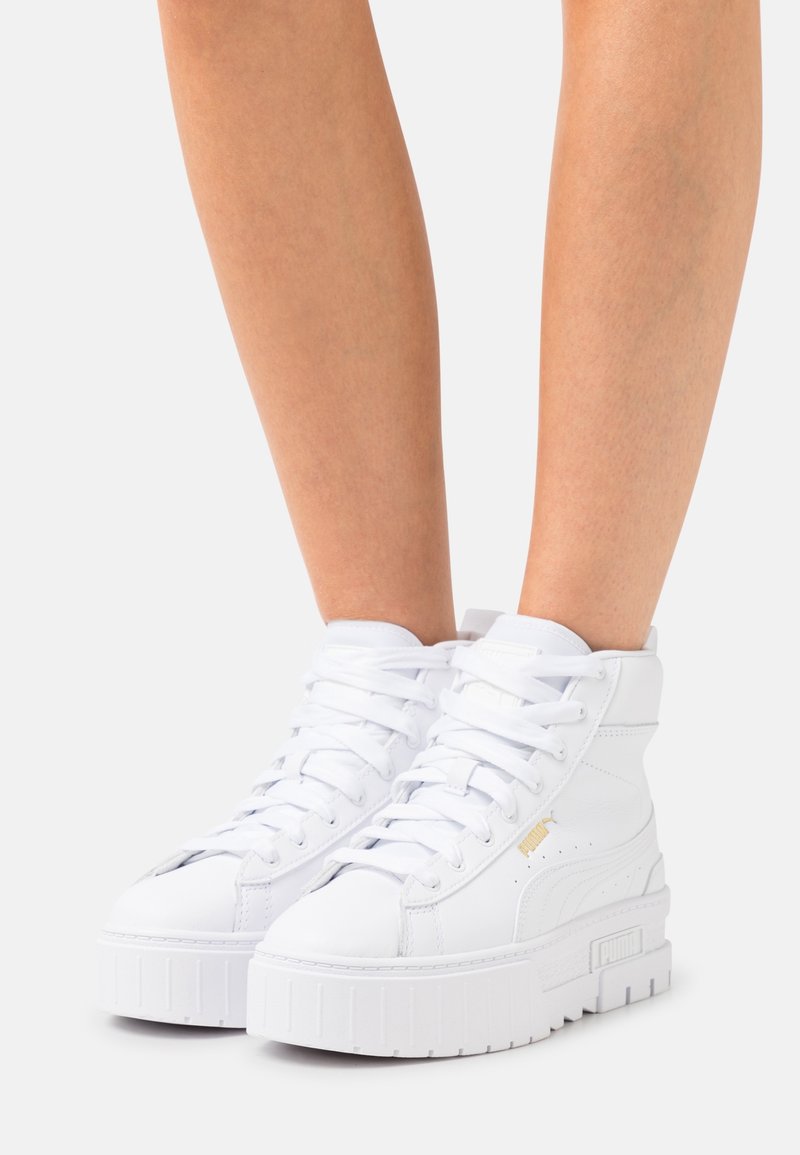 Jambes portant des baskets blanches à plateforme haute avec semelles épaisses et lacets sur un fond clair uni.