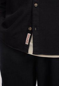 Camisa negra texturizada con cierre de botones, que presenta una pequeña etiqueta roja y blanca que dice "REVOLUCIÓN" en el dobladillo. Camiseta blanca visible por debajo.