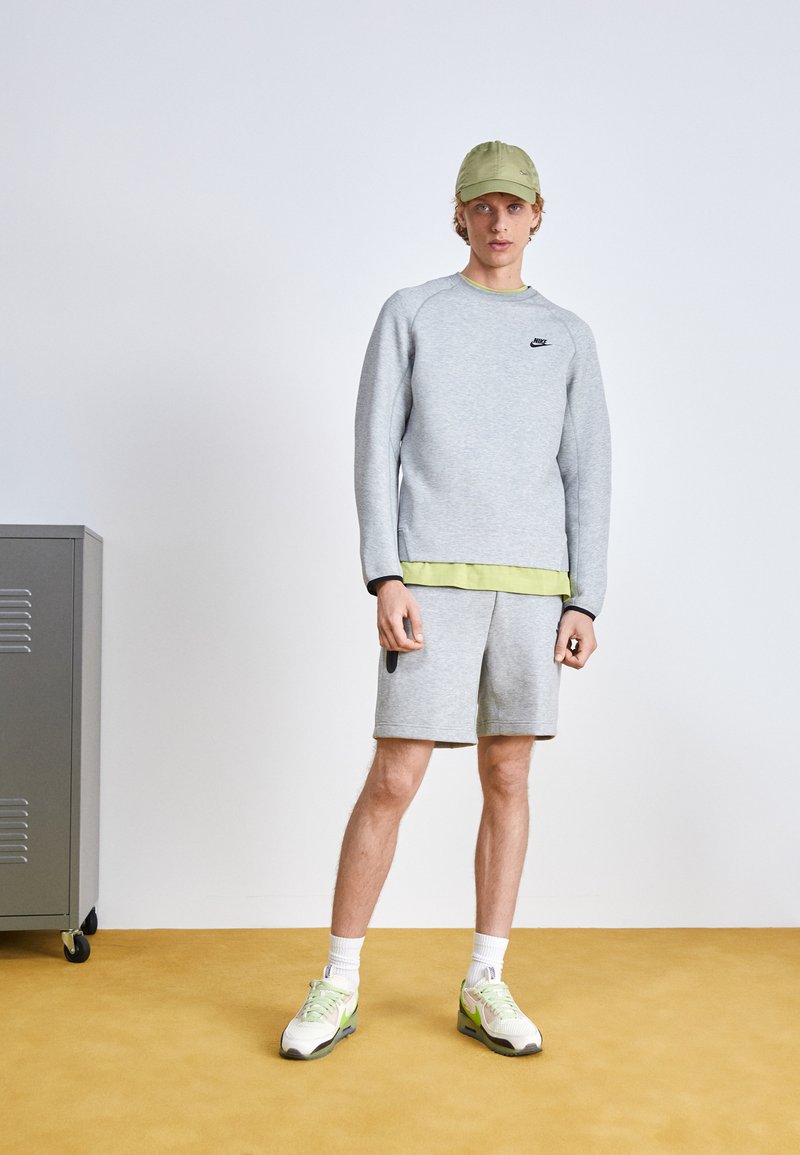 Un sweat-shirt gris clair avec un logo noir, associé à un short gris. Porté avec une casquette vert clair et des baskets blanches avec des accents néon.