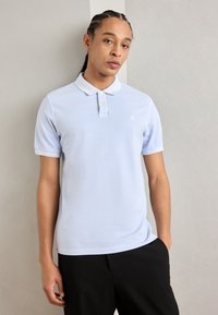 Lyseblå polo skjorte med en solid tekstur, tre-knaps placket og en liten logo på brystet. Stylet med svarte bukser.