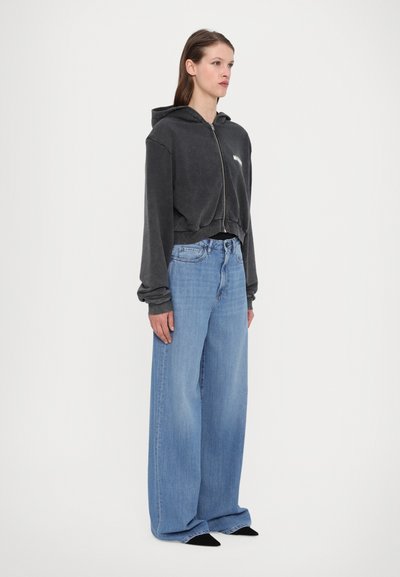 Gekürzter schwarzer Reißverschluss-Hoodie mit Kordelzug, kombiniert mit hellblauen weit geschnittenen Jeans. Der Stoff hat eine weiche, strukturierte Oberfläche.